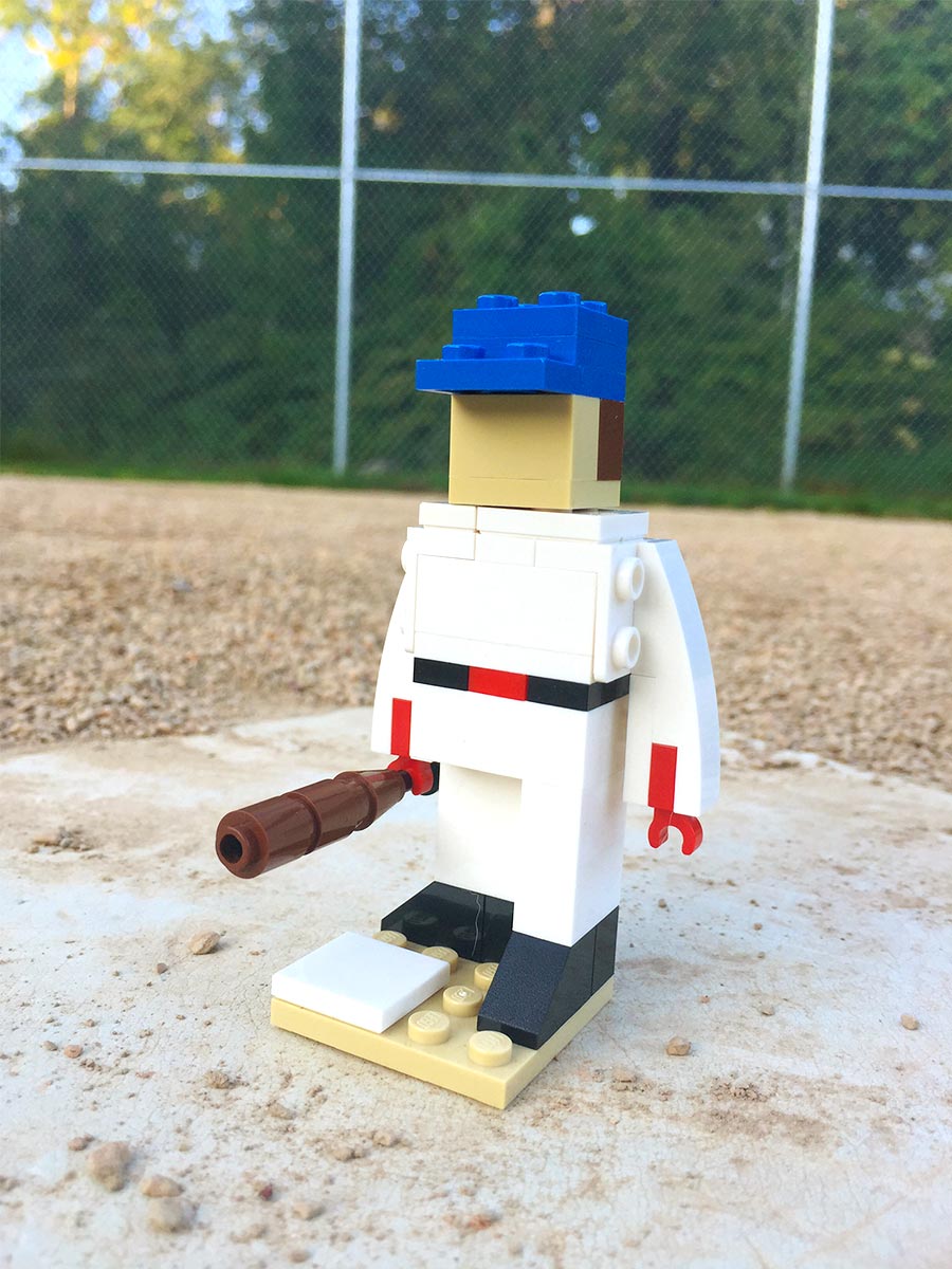 A LEGO baseball player with bat in hand, stands on the home plate of a baseball field. 